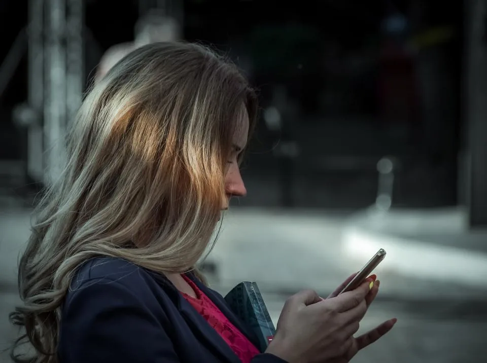 A young woman with long hair, in profile on a dim street, staring at her phone screen